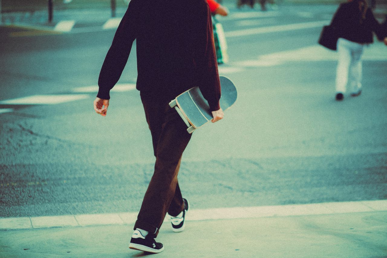 A skateboarder carrying a skateboard walks along a city street, capturing urban life.